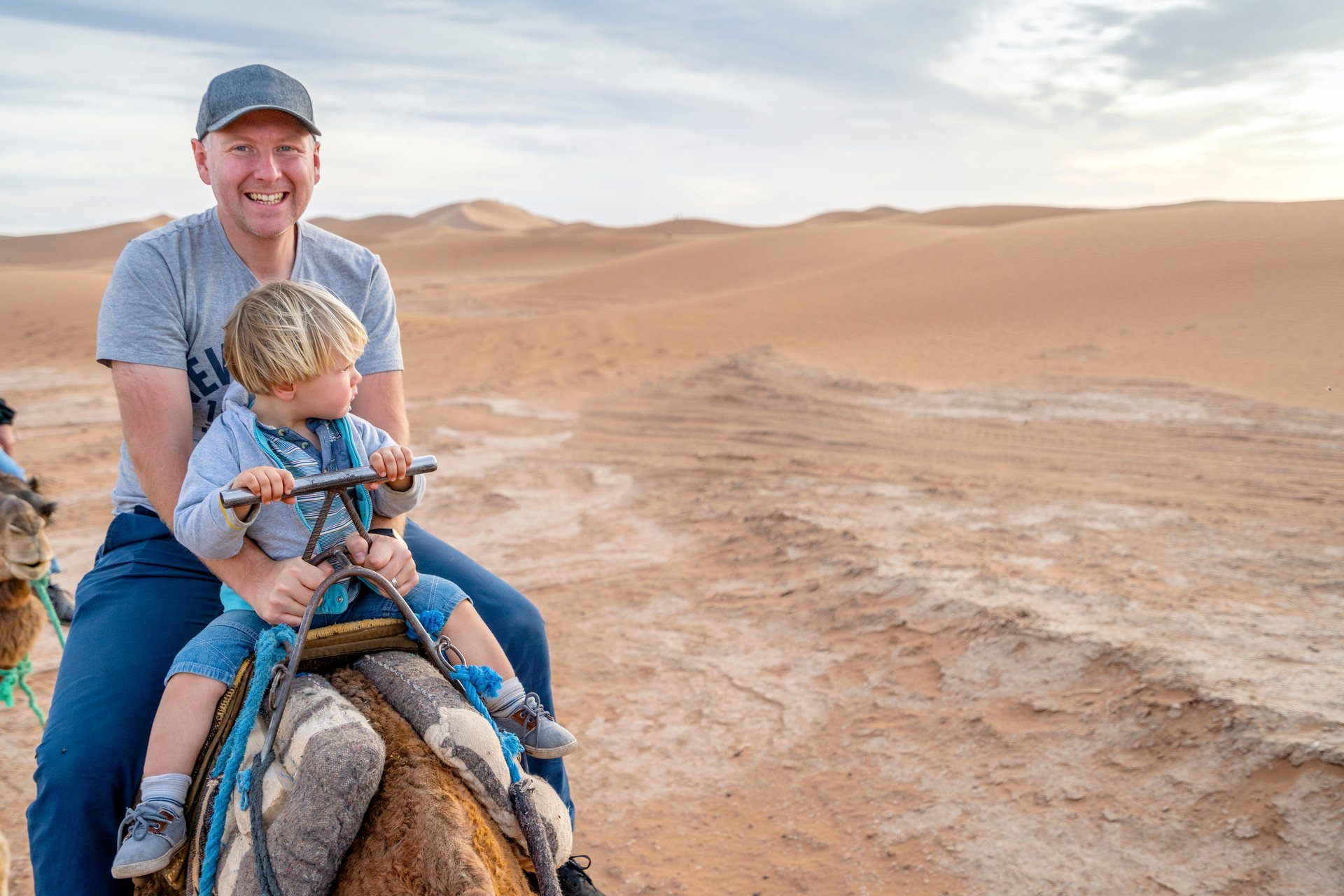 Vader en jong kind rijden samen op een kameel door de Sahara bij Merzouga, met de zandduinen van Erg Chebbi op de achtergrond in Marokko