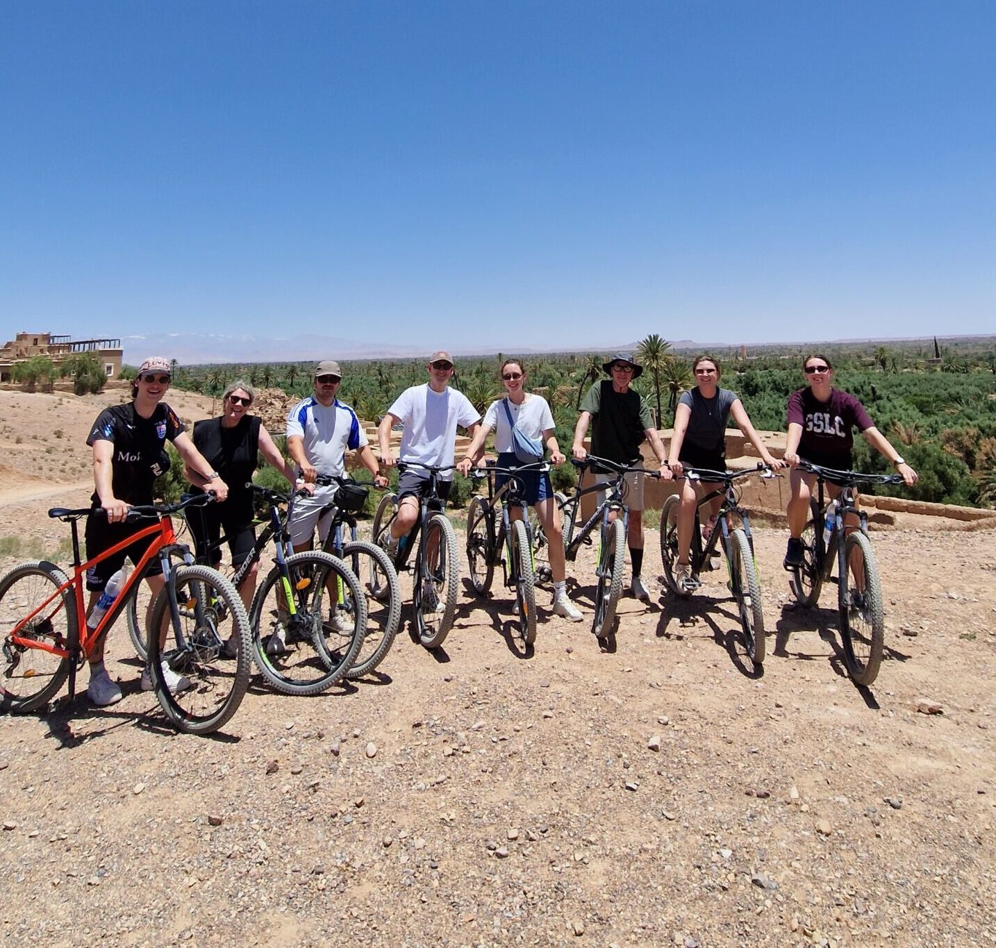 Groep fietsers poseert met mountainbikes op een rotsachtig plateau met uitzicht over een groene oase in Marokko.