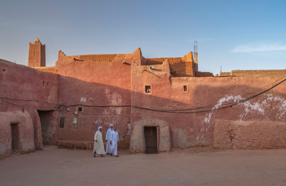 Rode lemen gebouwen in een ksar bij Timimoun met bewoners op het plein en een minaret op de achtergrond